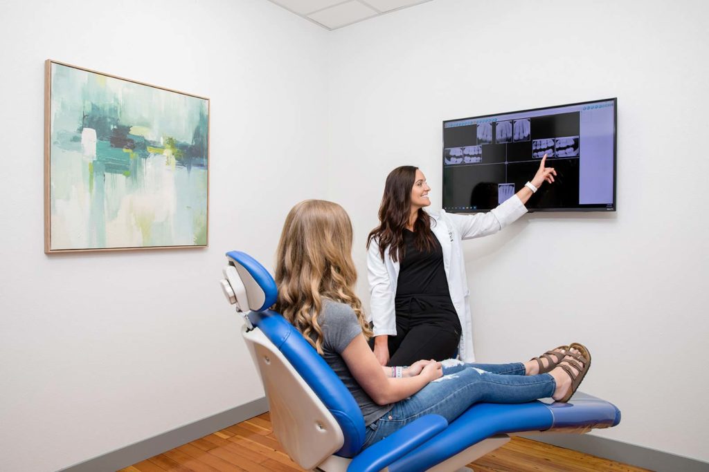 A dentist points to a dental x-ray shown on a tv while consulting with a dental patient who is seated in an exam chair