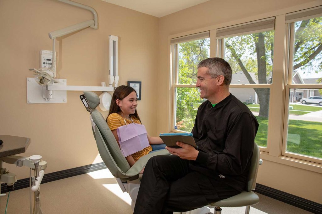 Dr. Burhoop is seated in a chair while speaking to a young dental patient who is seated in an exam chair