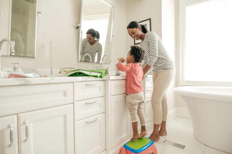 A child brushing their teeth with their mom in front of a sink and mirror