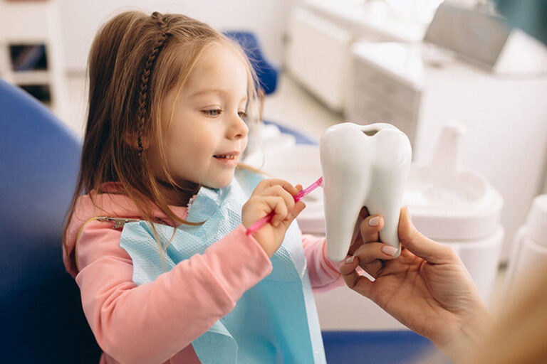 A pediatric dental patient sits in an exam chair and learns how to brush her teeth