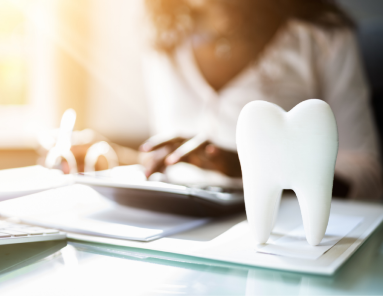 Close-up of a dental office desk with a model of a tooth in the foreground and a person working on documents in the background, representing financial planning and dental care.