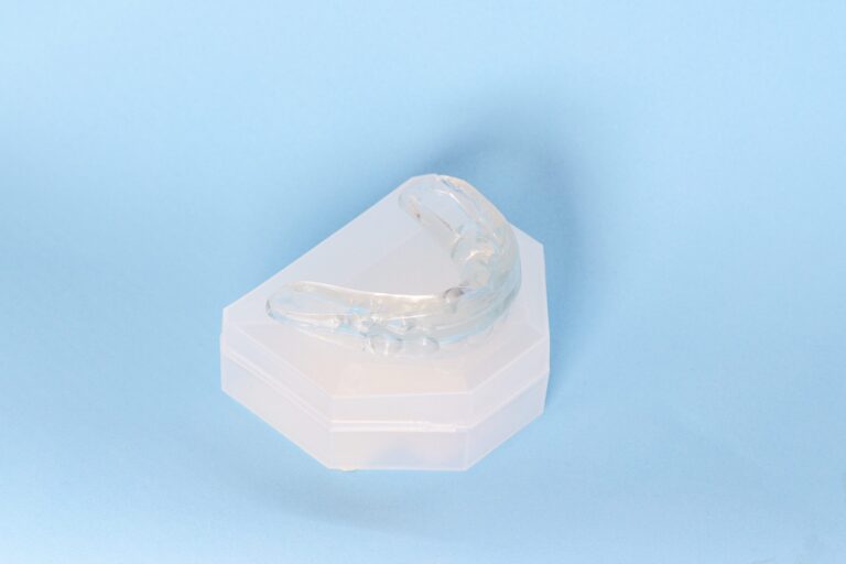 Transparent dental mouthguard rests atop a white hexagonal storage case against a light blue background. The appliance is custom-molded to fit upper teeth, typically used for bruxism, orthodontics, or sports protection.
