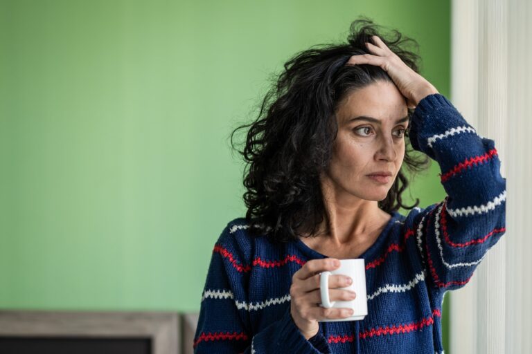 The image shows a person with curly hair wearing a blue sweater, holding a white mug in one hand and the other hand on their head, likely indicating stress.