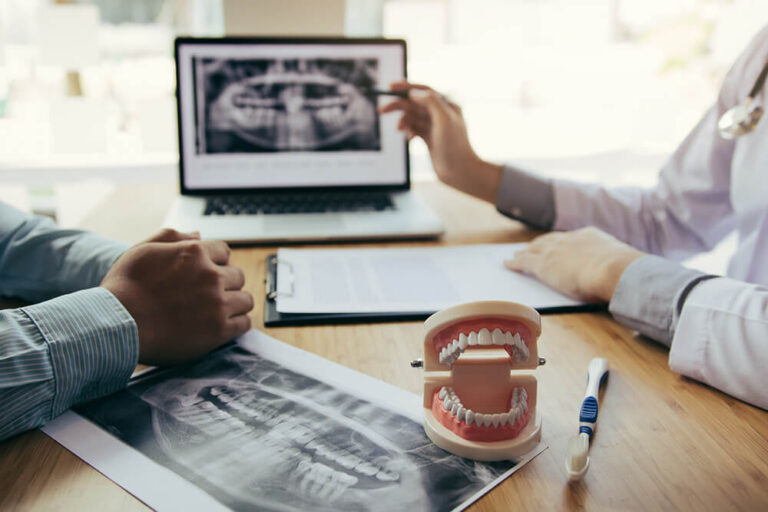 A dental consultation setting with a dentist and a patient discussing a treatment plan. The dentist is holding a pen and pointing at a document, while the patient has their hands clasped together. On the table, there is a clipboard with a dental X-ray image, a laptop displaying the same X-ray, a dental model, and a toothbrush.