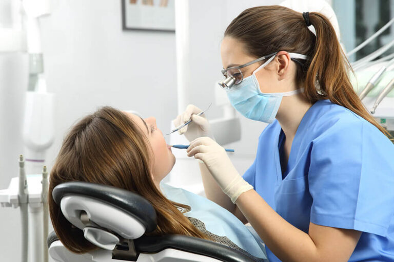 Dental professional in blue scrubs performing a procedure on a patient in a dental chair, with dental instruments and equipment visible in the background.