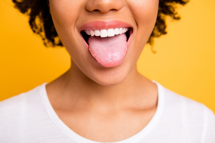A close-up of a woman's mouth smiling widely and sticking her tongue out against a bright yellow background.